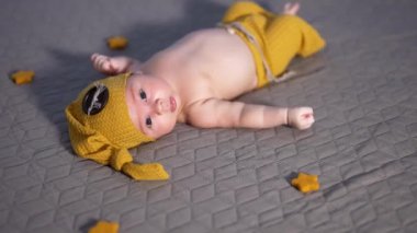 Sarı cüce kostümü giymiş sevimli bir erkek bebek yatakta yatıyor. Bebeğin etrafında dekoratif yıldızlar var. Bebek dili dışarı çıkarır..