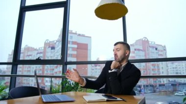 Bilgisayarda iş görüşmesi. Orta yaşlı girişimci büyük panoramik pencerelerin önündeki masada oturur. Arkaplanda şehir manzarası.