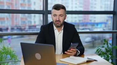 Ceketli ve takım elbiseli bir adam laptopta biriyle konuşuyor ve elinde akıllı telefon tutuyor. Ofisteki ortaklara video çağrısı. Görüşmeler dizüstü bilgisayarla yapılıyor.