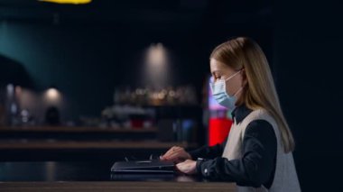 Maske takan sarışın kadın karanlık bir odada bilgisayarın başında çalışıyor. Geç saate kadar çalışan bir bayan. Yan görünüm.