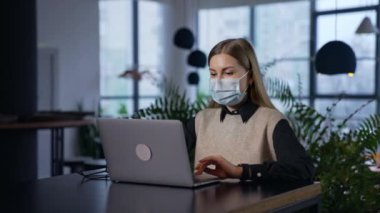 Laptopunda cerrahi maske takan bir kadın. Maskeli güzel bayan ofiste çalışıyor. Ofis işi karantina döneminde.