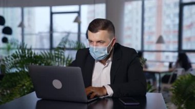 Cerrahi maske takan adam bir ofiste dizüstü bilgisayarın önünde çalışıyor. İşveren laptopunu kapatır, maskesini çıkarır ve geriye doğru fırlatır. Salgın zamanlarında ofis çalışması.