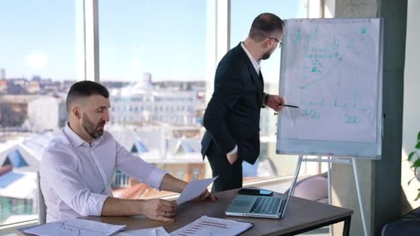 ライトオフィスで何かを議論する二人のビジネスマン 一人の男がテーブルに座って紙を持っている ホワイトボードに立ってチャートを見てコメントするビジネスパートナー — ストック動画
