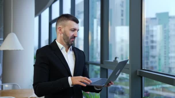 男性起業家が窓の前に立って 彼の前にラップトップを持っています ビジネスはコンピューターで話す ノートブックの前に話してジェスチャーする男 — ストック動画