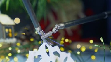 Robot kolların küçük pençeleri ipliği kağıt kar tanesine bağlıyor. Ekipmanların hassas hareketleri. Arkaplanda Noel süslemeleri.