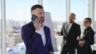 Duygusal iş adamı akıllı telefondan konuşuyor. İnsan aktif olarak konuşur ve hareket eder. Arkaplanda tabloda duran çalışanlar.