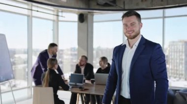 Takım elbiseli bir adam kameraya bakıp gülümsüyor. Genç bir iş adamının portresi. Arka plandaki masada iş birliği.