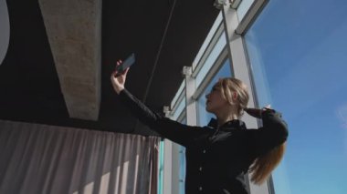 Önünde akıllı telefonu tutan çekici bir kadın var. Kadın güneşli bir ofiste kameranın önünde poz veriyor. Aşağıdan görüntüle.