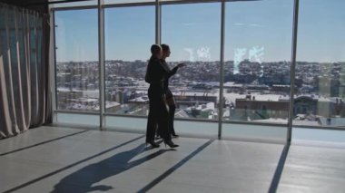 İki bayan iş arkadaşı panoramik pencereler boyunca gider. Bayan meslektaşlar geniş ofiste iletişim kuruyor. Arkaplanda şehir manzarası.
