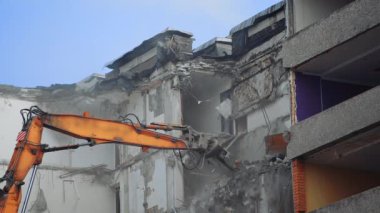 Güçlü kazıcı pençesi enkazı yere döküyor. Tozla kaplı inşaat alanı. Arkaplanda açık mavi gökyüzü.
