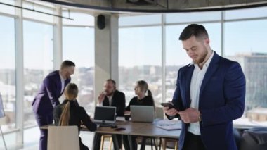 Erkek iş ortağı ofiste telefonunu kontrol ediyor. Çalışma arkadaşları işbirliği yapıyor ve arka planda tartışıyorlar..