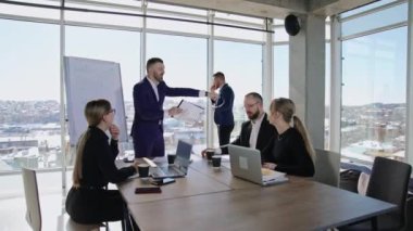 Meslektaşlar dizüstü bilgisayarların önünde masada oturup hoparlörleri dinliyorlar. Erkek ortak pencerede duruyor ve telefonda konuşuyor..