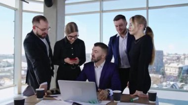 Erkek lider, takım arkadaşlarıyla birlikte dizüstü bilgisayarın önünde oturuyor. İş adamları iş yerinde iletişim kuruyor. Ofis pencerelerinde şehir manzarası.