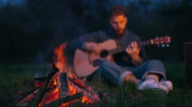 Adam ateşin yanında oturup gitar çalıyor. Gitarlı bir adam geceleri dışarıda şenlik ateşinin yanında dinleniyor.