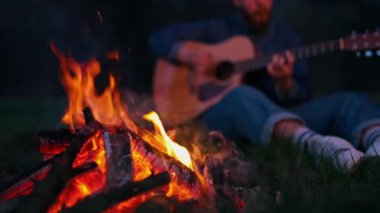 Gitarlı şenlik ateşi başında oturan adam. Yalnız bir adam ormanın yanındaki sahilde şenlik ateşinde oturmuş akustik gitar çalıyor.