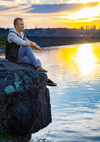 Man Contemplating Nature\'s Beauty. A man sitting on top of a rock next to a body of water