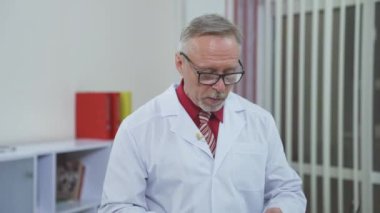 Uzaktan kumandalı hasta çevrimiçi. Beyaz önlüklü kıdemli erkek doktor görüntülü konuşmada telefonla konuşuyor. Deneyimli doktor, klinikteki web kamerasıyla tedaviyi açıklıyor.