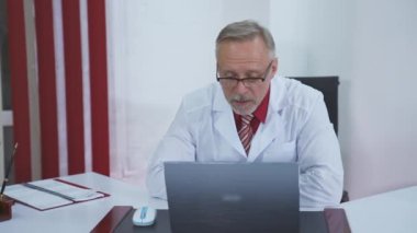 Profesyonel doktor uzak bir hastayla konuşur. Klinikte dizüstü bilgisayara bakarken görüntülü konuşma yapan beyaz üniformalı bir erkek doktor..