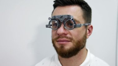 Test gözlüklü genç sakallı bir adamın portresi. Hasta, klinikte görme yeteneğini optik deneme çerçevesiyle inceliyor. Oftalmoloji kavramı.