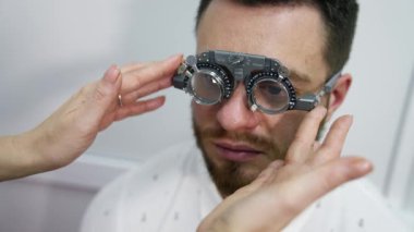 Doktor optik deneme çerçevesi ile insanın görüşünü kontrol eder. Görme yeteneğini göz doktorunda test eden genç bir erkek hastanın portresi..