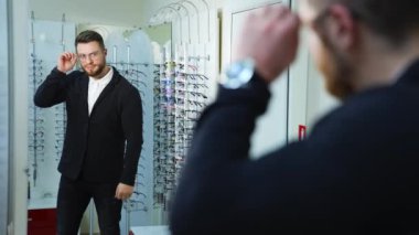 Optik dükkanında yeni gözlük deneyen bir adam. Yakışıklı adam yeni gözlük takıyor ve aynadaki yansımasına bakıp gülümsüyor. Göz giyimi konsepti.