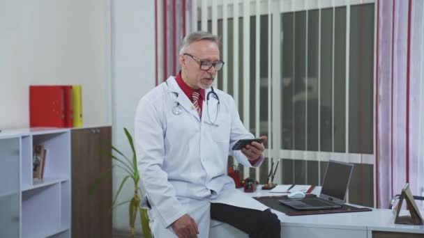 检疫期间的医疗咨询 上了年纪的医生 戴着眼镜坐在橱柜里 用电话与病人进行视频通话 — 图库视频影像