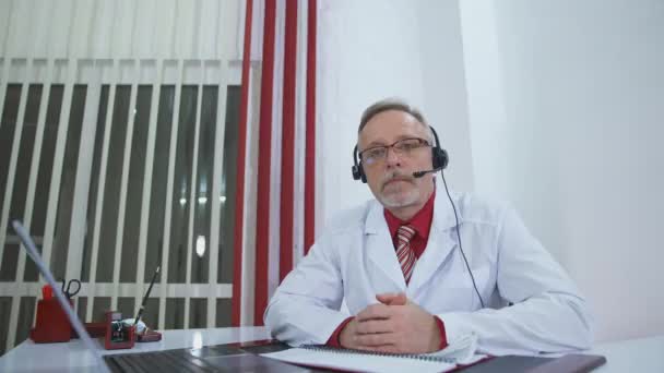 高齢の医師は ビデオ通話中に患者を診察する 医療ユニフォームとヘッドセットのシニア男性医師は 遠隔相談を受けています テレメディシンコンセプト — ストック動画