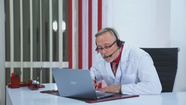 遠くの患者と話している男性医師 ヘッドセットのシニアドクターは 医療キャビネットのラップトップでビデオチャットを使用して患者にリストします — ストック動画