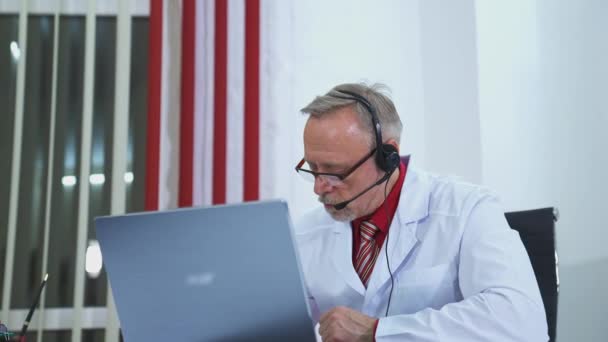 テレメディシン 古い医師はラップトップから患者を診察する 男性の医者はヘッドセットを身に着け 診療所の患者にオンラインビデオ通話相談をする — ストック動画