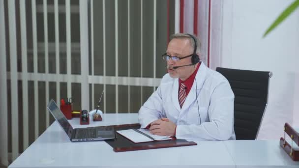 远程医疗咨询 穿着白色医疗外套和耳机的专业男医生在笔记本电脑上进行电话会议 医生用网络摄像头解释治疗 — 图库视频影像