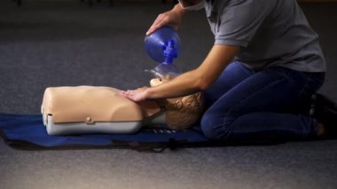 Eğitmen bir manken üzerinde nefes egzersizi gösteriyor. Paspasın üstünde yatan bir kadın mankenin üzerinde tıbbi eğitim veriyor..