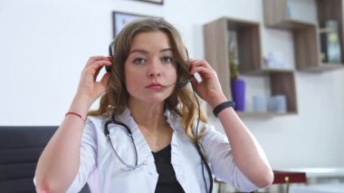 Kulaklık takmış genç bir doktorun portresi. Beyaz önlüklü kadın bir tıp asistanı uzaktaki bir hastayla görüntülü konuşma yapmak için kulaklık takıyor. Teletıp, uzaktan sağlık hizmeti kavramı.