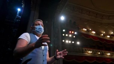 Tıbbi maskeli aktör konuşuyor. Sahnede sahne ışıkları önünde duran ve salgın sırasında konuşan bir adam. Aşağıdan görüntüle.