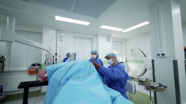 Modern ameliyathane. Mavi üniformalı tıbbi personel cerrahi ekipmanların arka planında bir hastayı ameliyat ediyor. Ameliyat masasında baygın bir hasta var..