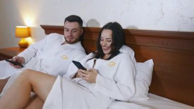 Yatakta beyaz elbiseli güzel bir çift. Çekici bir kadın, bir otelde yatakta yatarken kocasıyla selfie çektiriyor. İyi tatiller..