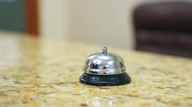 Otel zili resepsiyon masasında. Adamın eli oteldeki resepsiyonisti aramak için zil tuşuna basıyor. Müşteri metal alarma dokunuyor. Yakın plan..