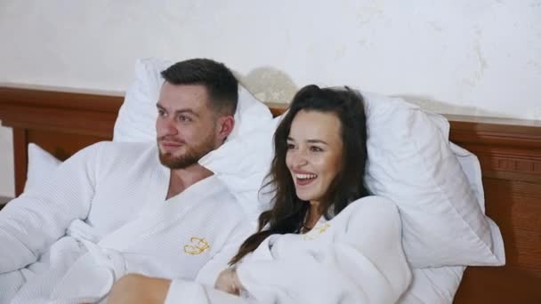 可爱的夫妇在酒店的床上 迷人的年轻女子和身穿白衣的英俊男子躺在床上 一起看电视 — 图库视频影像