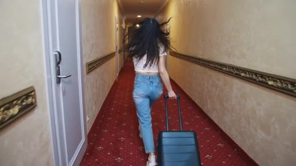 屋内スーツケース付きのハッピー女性旅行者 トロリーバッグでホテルの廊下を歩き 喜んでジャンプするカジュアルな服を着た若い女性 バケーションコンセプト — ストック動画
