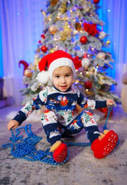 Büyüleyici bir görüntü, Noel Baba şapkasıyla süslenmiş, göz kamaştırıcı bir Noel ağacının önünde neşeli bir şekilde duran sevimli bir bebeği yakalıyor..