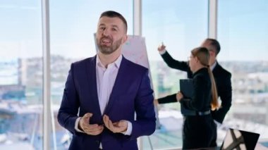 Kostüm giymiş girişimci şirketinden bahsediyor. Başarılı iş adamı, ofiste çalışan meslektaşlarının arka planında yer alan yeni bir proje hakkında konuşuyor..