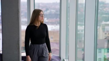Modern ofiste kendine güvenen bir iş kadını. Gözlüklü ve şık elbiseli güzel bir kız büyük ofis pencerelerinin yanında duruyor ve şehir manzarasına bakıyor..