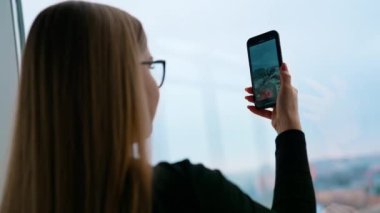 Kapalı mekanda cep telefonu olan bir kız. Ofis penceresinden şehir manzarasının fotoğrafını çeken genç bir kadın. Yakın plan..
