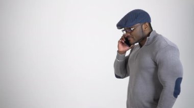 Stüdyoda telefonu olan ciddi bir zenci. Gri kazaklı ve mavi şapkalı Afro-Amerikan adam beyaz arka planda cep telefonuyla konuşuyor. Yan görünüm.