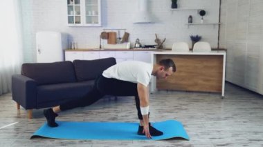 Genç adam evde fitness yapıyor. Tişörtlü ve pantolonlu bir adam evde, geniş mutfağında paspas üzerinde egzersiz yapıyor..