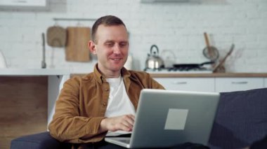 Genç adam mutfakta dizüstü bilgisayar kullanıyor. Laptop aracılığıyla sohbet eden mutlu bir adam. İnternette evden çalışıyor. Coronavirus karantinası sırasında evden uzak çalışma.