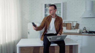 Evden uzak bir iş. Genç adam dizüstü bilgisayarla masada oturuyor ve mutfak arka planındaki serbest iş sırasında akıllı telefon kullanıyor..