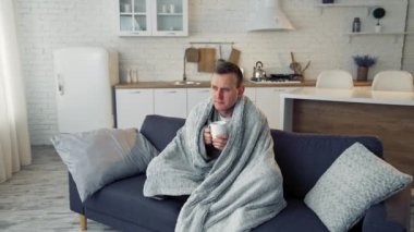 Evde sıcak içecek içen kötü bir adam. Mutfaktaki kanepede sıcak battaniyeyle kaplanmış hasta genç bir adam oturuyor. Koronavirüs salgını.