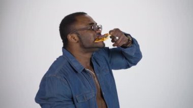 Gözlüklü siyah adam pizza yiyor. Mavi gömlekli Afro-Amerikalı adam beyaz stüdyo arka planında lezzetli bir pizza tadı alıyor. Profil görünümü.
