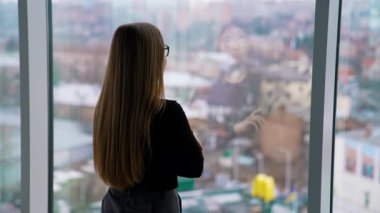 Camda duran gözlüklü çekici iş kadını. Uzun saçlı kadın şehir manzarasında pencereden dışarı bakıyor. Arka görünüm.