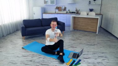 Genç adam evde egzersiz yapıyor. Adam yerde bir paspasın üzerinde oturuyor ve biberonla dengesini koruyor. Karantina sırasında evde spor.